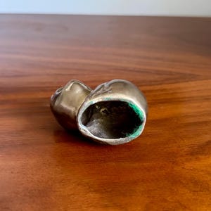May include: A small, metallic, antique-looking object, possibly a decorative piece or a small container. The object has a rounded shape with a hollow interior lined with green fabric. The exterior has a tarnished, aged appearance.