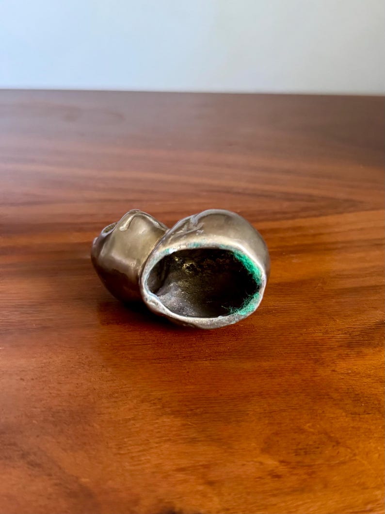 May include: A small, metallic, antique-looking object, possibly a decorative piece or a small container. The object has a rounded shape with a hollow interior lined with green fabric. The exterior has a tarnished, aged appearance.