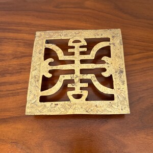 May include: A square, gold-coloured metal decorative piece with an intricate, openwork design. The design features a central, stylized symbol within a square frame, with floral patterns along the edges. The piece is set against a dark wood surface.