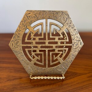 Puede incluir: Un salvamanteles hexagonal dorado con un intrincado diseño calado, un símbolo central y un borde floral. El salvamanteles tiene una base de alambre retorcido, probablemente para platos calientes.