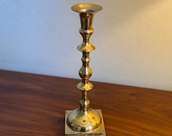 Puede incluir: Un candelero de latón con base cuadrada y un tallo decorativo escalonado. El candelero es de color dorado cálido y está diseñado para sostener una sola vela. El diseño general es clásico y elegante.