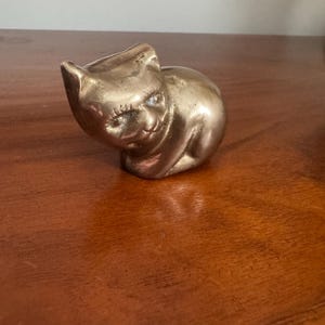 May include: A small, brass cat figurine with a smooth, polished surface. The cat is in a seated position with its head tilted slightly to the side. The figurine has detailed features, including ears, eyes, and a smiling expression. The cat is a decorative item.
