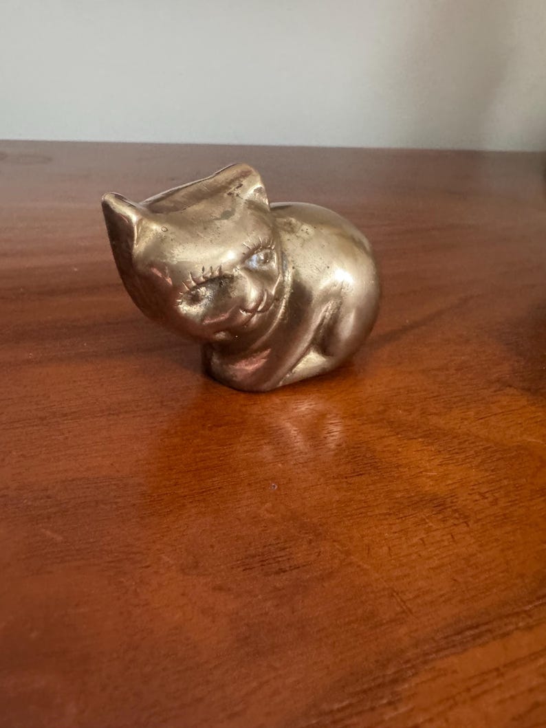 May include: A small, brass cat figurine with a smooth, polished surface. The cat is in a seated position with its head tilted slightly to the side. The figurine has detailed features, including ears, eyes, and a smiling expression. The cat is a decorative item.