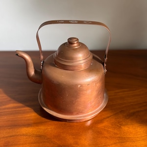May include: A copper-colored teapot with a curved spout, a domed lid, and a handle. The teapot sits on a small, round base. The handle has decorative markings. The teapot is on a wooden surface.