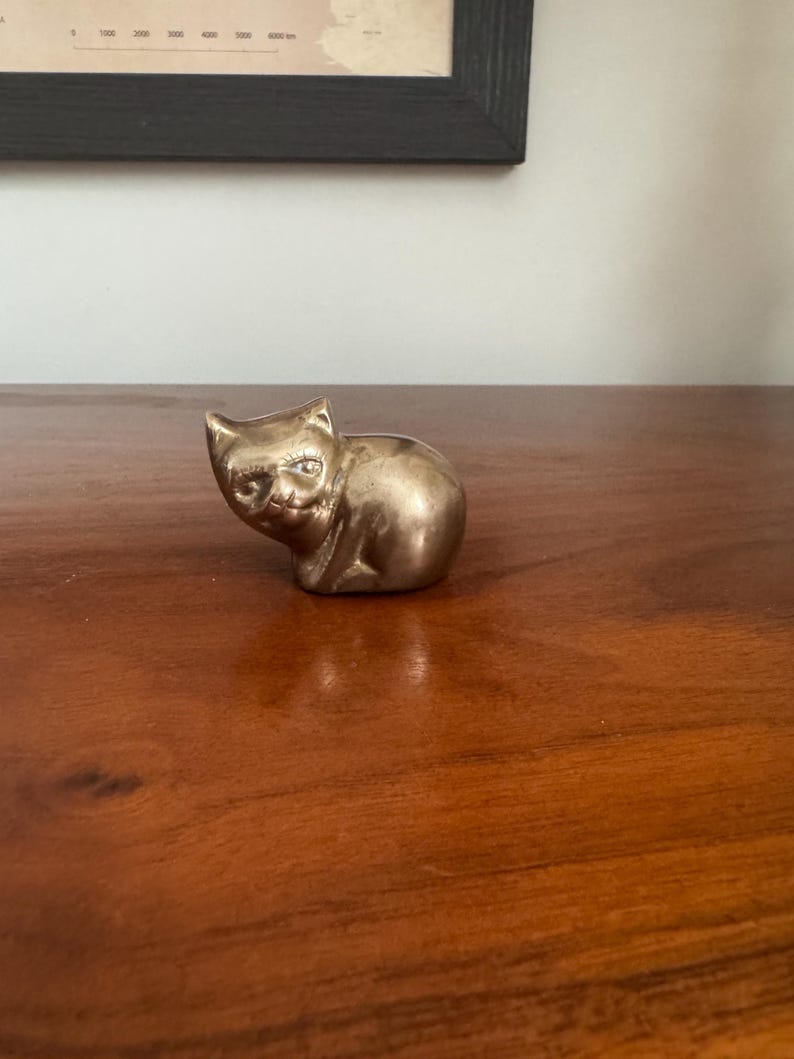 May include: A small, brass cat figurine with a seated pose. The cat has detailed facial features and a smooth, polished surface. The figurine is placed on a wooden surface, with a framed map in the background.