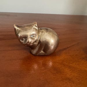 May include: A small, brass cat figurine in a seated position. The cat has detailed facial features, including eyes and a nose. The brass has a warm, golden hue, and the figurine is placed on a wooden surface.