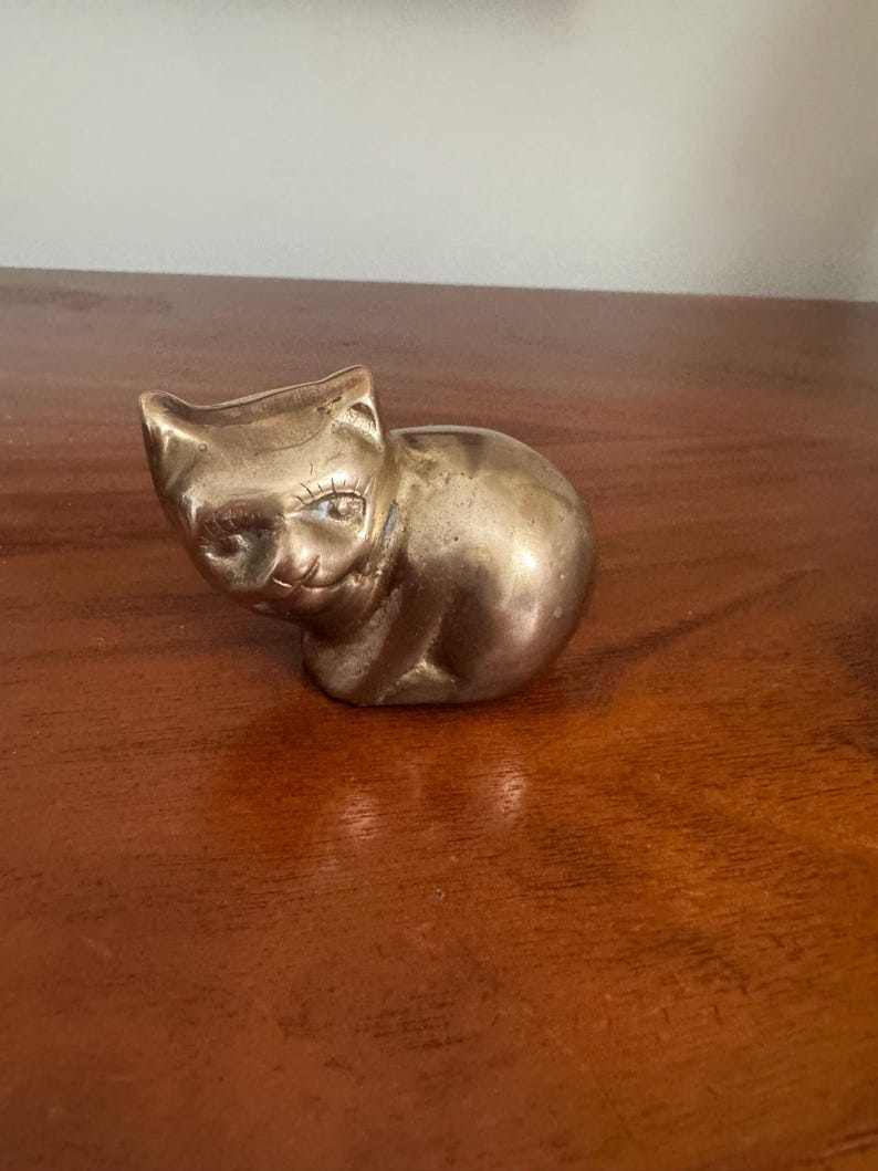 May include: A small, brass cat figurine in a seated position. The cat has detailed facial features, including eyes and a nose. The brass has a warm, golden hue, and the figurine is placed on a wooden surface.