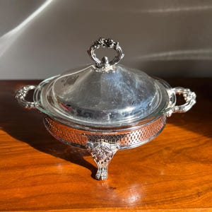 Könnte beinhalten: Eine silberbeschichtete Servierschale mit Glaseinsatz und gewölbtem Deckel. Die Schale hat verzierte Griffe und Füße sowie einen dekorativen Griff am Deckel. Die Schale steht auf einer Holzoberfläche.