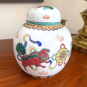 Peut inclure: Un pot en céramique blanc avec un couvercle, orné d'illustrations colorées. Le pot présente une créature mythique rouge et verte, une roue jaune et d'autres éléments décoratifs. Le couvercle a un design assorti.