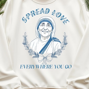 Può includere: Felpa bianca con una grafica blu di Madre Teresa con una ghirlanda di fiori intorno a lei. Il testo "Spread Love Everywhere You Go" è stampato sotto l'immagine.