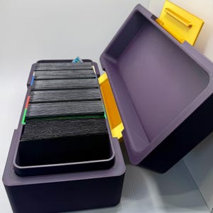 May include: A purple card storage box with a yellow lid. The box is open and filled with black cards. The box has a colorful stripe on the side.