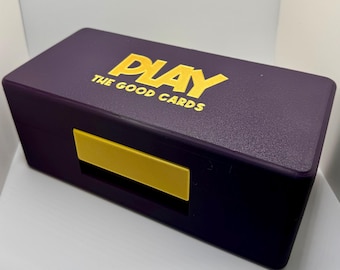 Op de afbeelding: Een paarse doos met een gouden "PLAY THE GOOD CARDS" logo. De doos heeft een gele sluiting.