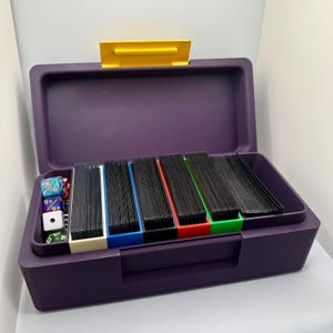May include: A purple plastic storage case with a yellow lid. The case is open and contains five stacks of black cards, each separated by a different colored divider: white, blue, red, and green. There are also several dice in the case.