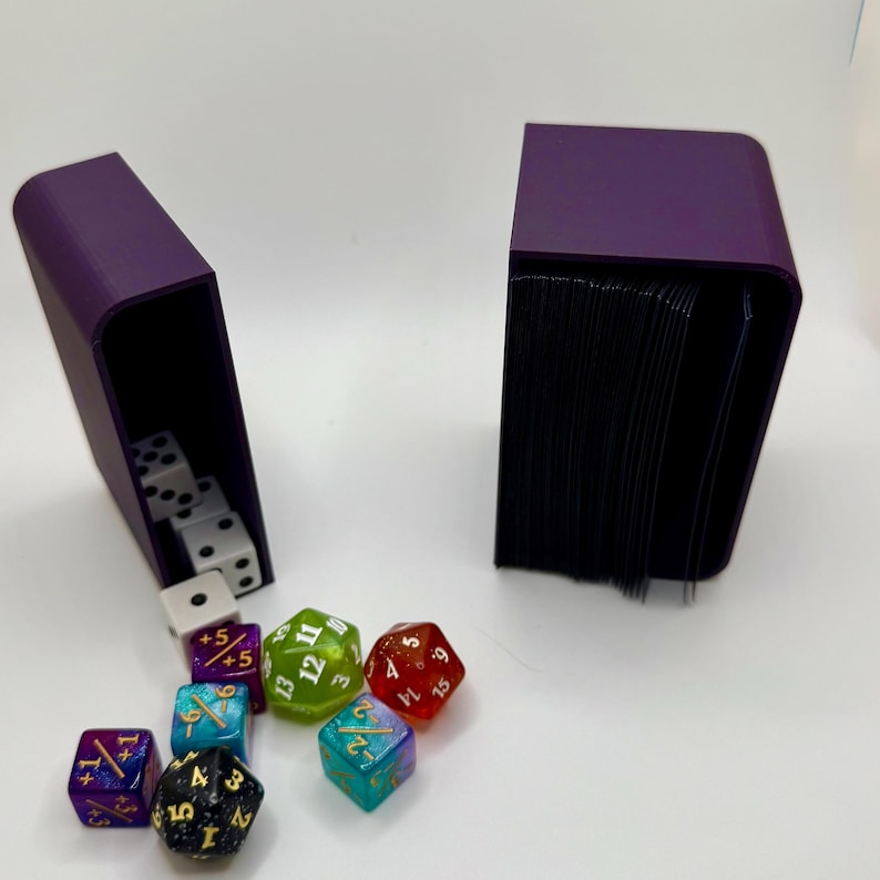 May include: Two purple boxes, one with a stack of cards and the other with dice. The dice are multi-colored and have different numbers on them. The dice are scattered on a white surface.