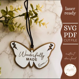 May include: A wooden butterfly decoration with a white centre and brown edges, inscribed with "Wonderfully Made" and the name "Rowan." It has a black hanging string and floral embellishments. The words "laser ready," "SVG PDF CUT FILE," and "instant download" are also visible.