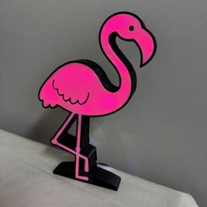 May include: A pink flamingo-shaped decorative object with a black outline. The flamingo is standing on one leg, with its head and neck curved. The object is made of a bright pink material and has a black base.
