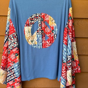 May include: A light blue long-sleeve shirt featuring a large circular peace sign applique on the chest. The peace sign and the long sleeves are constructed from a patchwork fabric pattern containing red, yellow, blue, and white floral motifs.