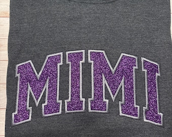 May include: A dark charcoal gray crewneck sweatshirt featuring the word MIMI embroidered across the chest in a bold, arched collegiate font. The lettering is filled with a vibrant purple glitter texture and outlined with a thin silver gray border.