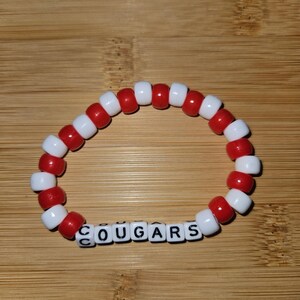 May include: Red and white beaded bracelet with the word "COUGARS" spelled out in black letter beads.