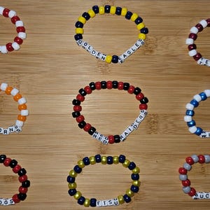 May include: Nine colorful beaded bracelets with white letter beads spelling out team names, including "Cougar", "Golden Eagles", "Texas A&M", "Longhorns", "Red Raiders", "Wildcats", "Bulldogs", "ETSU", and "Buckeyes".