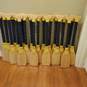 May include: A group of wooden paddles with black and yellow rope handles. The paddles are arranged in a row, standing upright.