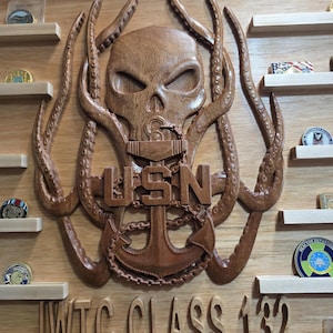 Puede incluir: Placa de madera con un diseño tallado de una calavera con tentáculos, un ancla y las letras "USN". También está presente el texto "WTC CLASS 132". Pequeñas estanterías exhiben monedas. La madera tiene un acabado natural.