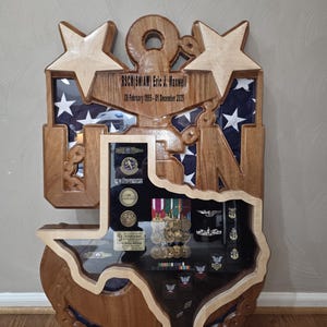Könnte beinhalten: Eine Holzvitrine in Form eines Ankers und des Bundesstaates Texas. Die Vitrine enthält Militärmedaillen und Erinnerungsstücke. Die Oberseite der Vitrine hat zwei Sterne und den Text "RSMC(SW/AW) Eric J. Maxwell 08 February 1995 - 01 December 2025".