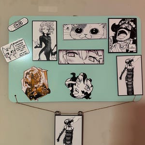 A grid arrangement of eight magnets and one orange/black sticker displayed on a stainless steel refrigerator. The collection features black and white manga panels of the Bomb Devil (Reze), Gol D. Roger, Tatsumaki, a cat face, a pair of eyes with "ARA ARA," a 'Boykisser License' meme, and a character with the quote, "LET ME SEE YOUR POTENTIAL."