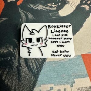 The Boykisser License charm resting on a Japanese-style desk mat featuring a large black cat and a city/mountain scene. This image showcases the front of the license.