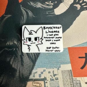 The Boykisser License charm resting on a Japanese-style desk mat featuring a large black cat and a city/mountain scene. This image showcases the front of the license.