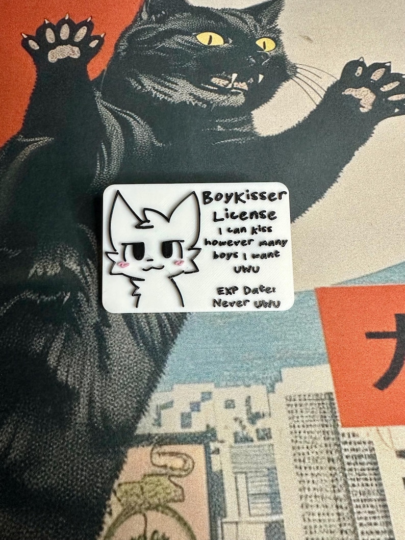 The Boykisser License charm resting on a Japanese-style desk mat featuring a large black cat and a city/mountain scene. This image showcases the front of the license.