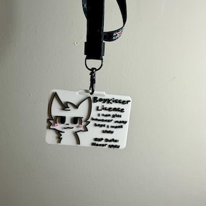 The Boykisser License attached to a black lanyard with a clip, hanging against a light wall, demonstrating the item's use as a badge or lanyard attachment.