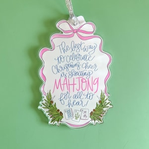 May include: A clear acrylic Christmas ornament with a pink bow and border. The ornament reads "The best way to celebrate Christmas cheer is shouting MAHJONG for all to hear!" with Mahjong tiles and greenery.