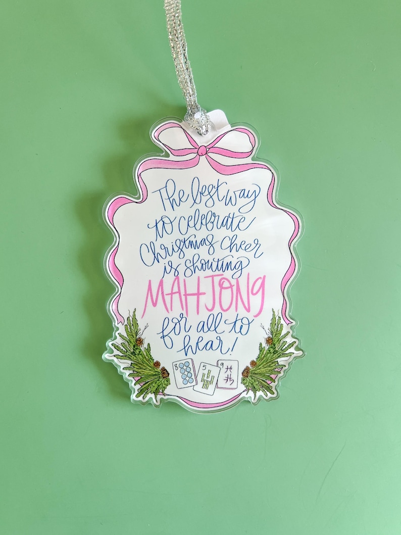May include: A clear acrylic Christmas ornament with a pink bow and border. The ornament reads "The best way to celebrate Christmas cheer is shouting MAHJONG for all to hear!" with Mahjong tiles and greenery.