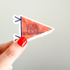 May include: A red and white pennant sticker with a black outline. The sticker features a line drawing of a building with a dome, surrounded by trees. The text "The University of North Carolina" is written in black at the bottom of the sticker.