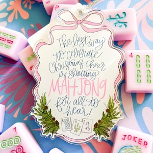 May include: A festive Christmas ornament with a pink bow and the text "The best way to celebrate Christmas cheer is shouting MAHJONG for all to hear!" surrounded by pink Mahjong tiles. The tiles are pink with green and blue markings.