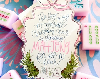 May include: A festive Christmas ornament with a pink bow and the text "The best way to celebrate Christmas cheer is shouting MAHJONG for all to hear!" surrounded by pink Mahjong tiles. The tiles are pink with green and blue markings.