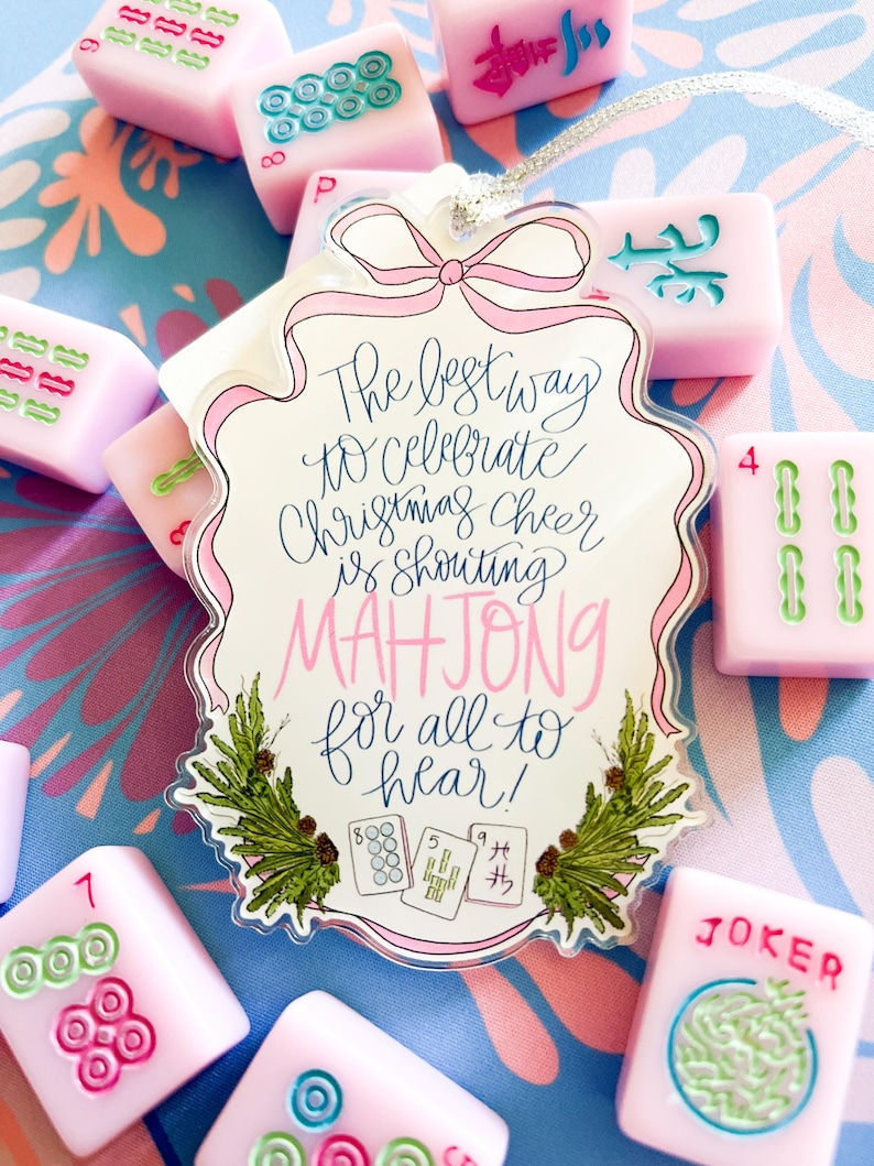 May include: A festive Christmas ornament with a pink bow and the text "The best way to celebrate Christmas cheer is shouting MAHJONG for all to hear!" surrounded by pink Mahjong tiles. The tiles are pink with green and blue markings.