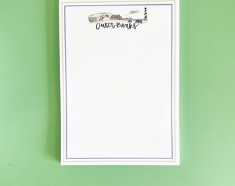 May include: A white notepad with a blue and gold border. The top of the notepad features a hand-drawn illustration of a lighthouse and the text "Outer Banks".