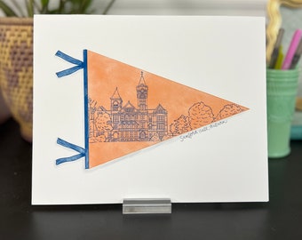 May include: A peach-colored pennant with a blue rope border and a blue ribbon at the top. The pennant features a line drawing of a building with a clock tower and the text "Samford Hall, Auburn".