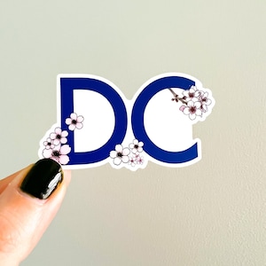 May include: A blue vinyl sticker with the letters "DC" in white, surrounded by pink cherry blossoms.