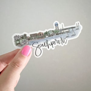 May include: A sticker with a watercolor illustration of a waterfront town with a lighthouse and boats. The text "Southport, NC" is written in cursive below the illustration.