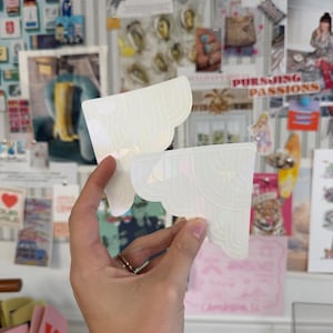 May include: Two iridescent white stickers with a scalloped edge are held up in front of a bulletin board. The stickers have a layered, wave-like design. The background includes various postcards, photos, and other decorative items.