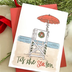 May include: A Christmas card with a beach theme. The card features a lifeguard stand with a red umbrella, a wreath, and the text "Tis the Season." The card is on top of a red envelope, with greenery and a red ribbon in the background.