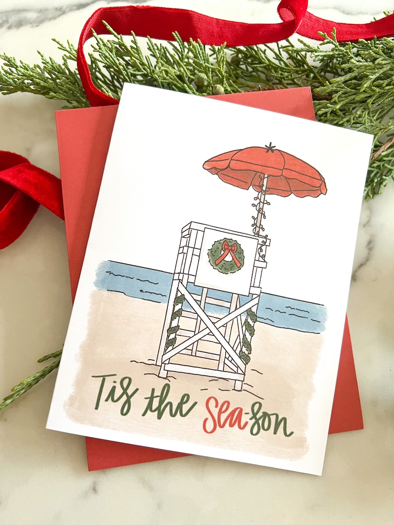 May include: A Christmas card with a beach theme. The card features a lifeguard stand with a red umbrella, a wreath, and the text "Tis the Season." The card is on top of a red envelope, with greenery and a red ribbon in the background.