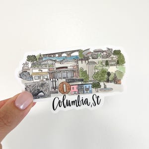 May include: A sticker featuring a watercolor illustration of Columbia, SC landmarks. The design includes a bridge, stadium, and various buildings, with the city name in cursive script. The sticker has a white background and is held by a hand.