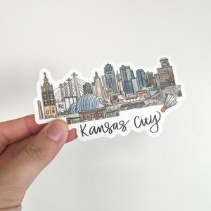 Puede incluir: Una pegatina con una ilustraci&oacute;n dibujada a mano del horizonte de Kansas City. El horizonte incluye edificios ic&oacute;nicos como el Country Club Plaza, el Power & Light District y el Sprint Center. La pegatina es blanca con letras negras que dicen "Kansas City".
