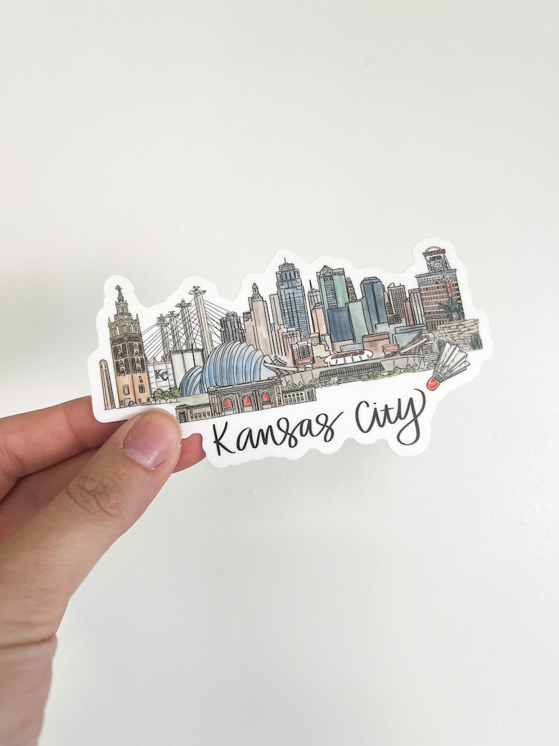 Puede incluir: Una pegatina con una ilustraci&oacute;n dibujada a mano del horizonte de Kansas City. El horizonte incluye edificios ic&oacute;nicos como el Country Club Plaza, el Power & Light District y el Sprint Center. La pegatina es blanca con letras negras que dicen "Kansas City".