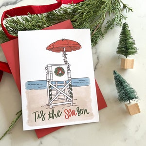 May include: A Christmas card with a beach theme. The card features a lifeguard stand with a red umbrella, a wreath, and the text "Tis the Season." The card is on a red envelope, with small decorative Christmas trees.