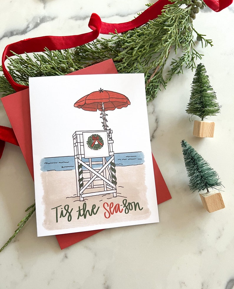 May include: A Christmas card with a beach theme. The card features a lifeguard stand with a red umbrella, a wreath, and the text "Tis the Season." The card is on a red envelope, with small decorative Christmas trees.